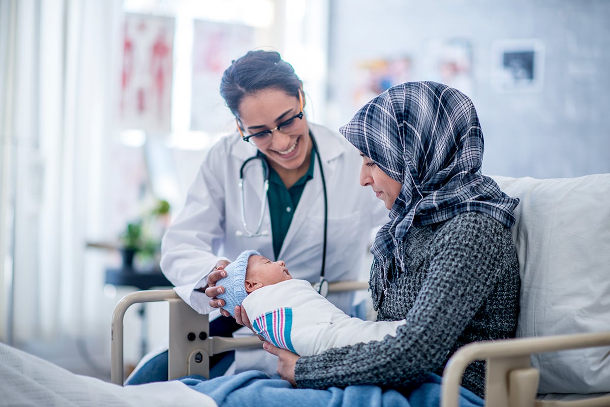 A mother and her newborn baby boy are indoors in a hospital. The mother is holding her baby while laying in the hospital bed. Their doctor is smiling down at the baby.